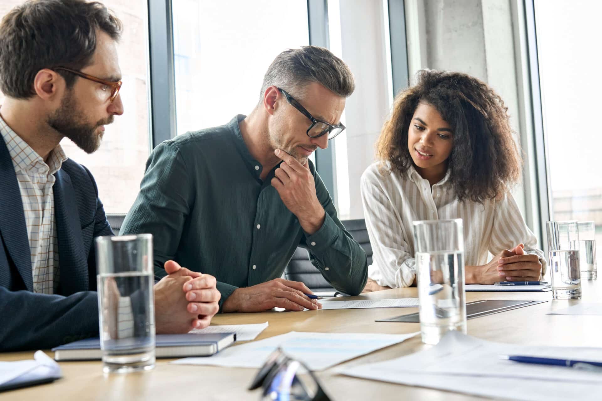 Focused doubtful mature businessman reading contract document thinking considering risks with professional lawyers legal experts executive team analyzing financial report sitting at office table.