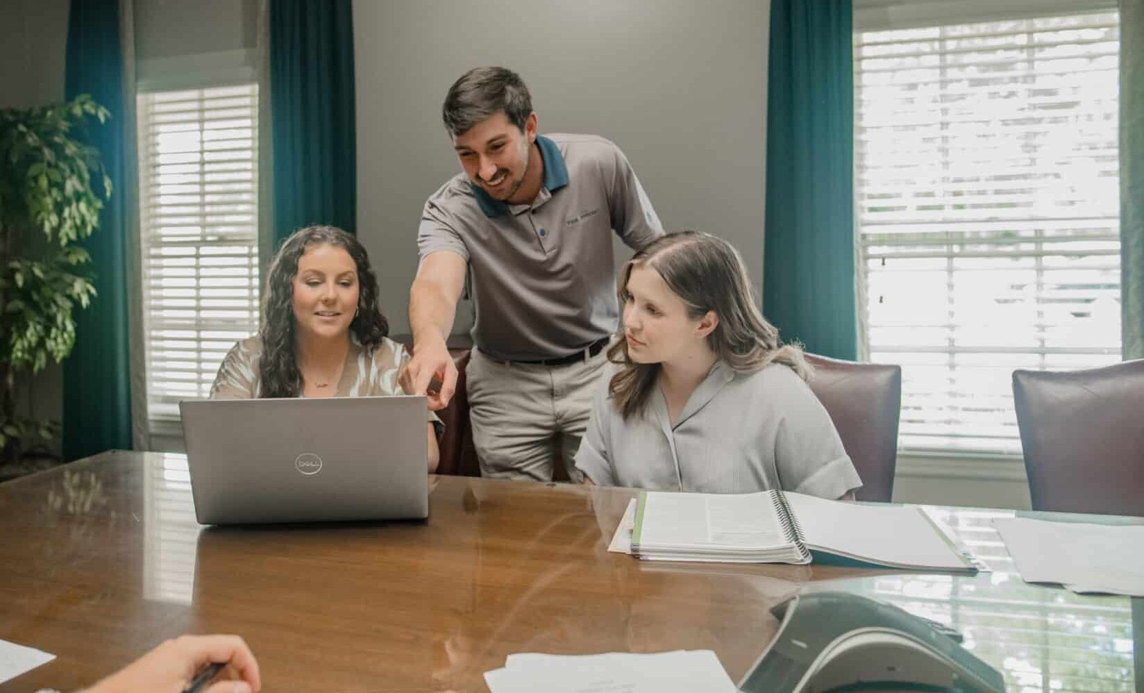 Three people are sitting around a wooden table with a laptop and documents. One person is standing and pointing at the laptop screen, while the other two are seated and focusing on the screen. The room has large windows with white blinds and green curtains.