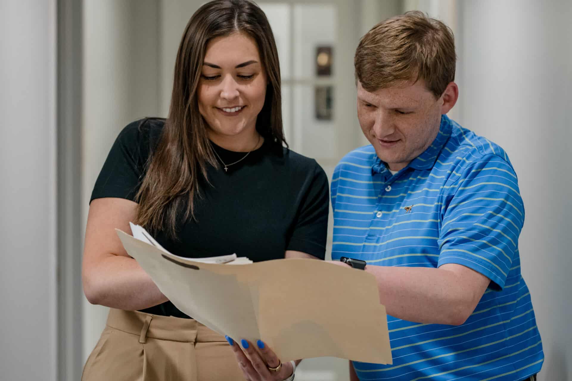 Two colleagues stand together in a hallway, smiling and reviewing documents. The woman, with long brown hair, is dressed in a black top and light-colored pants, while the man, with short red hair, wears a blue striped polo. Both appear engaged in their discussion.