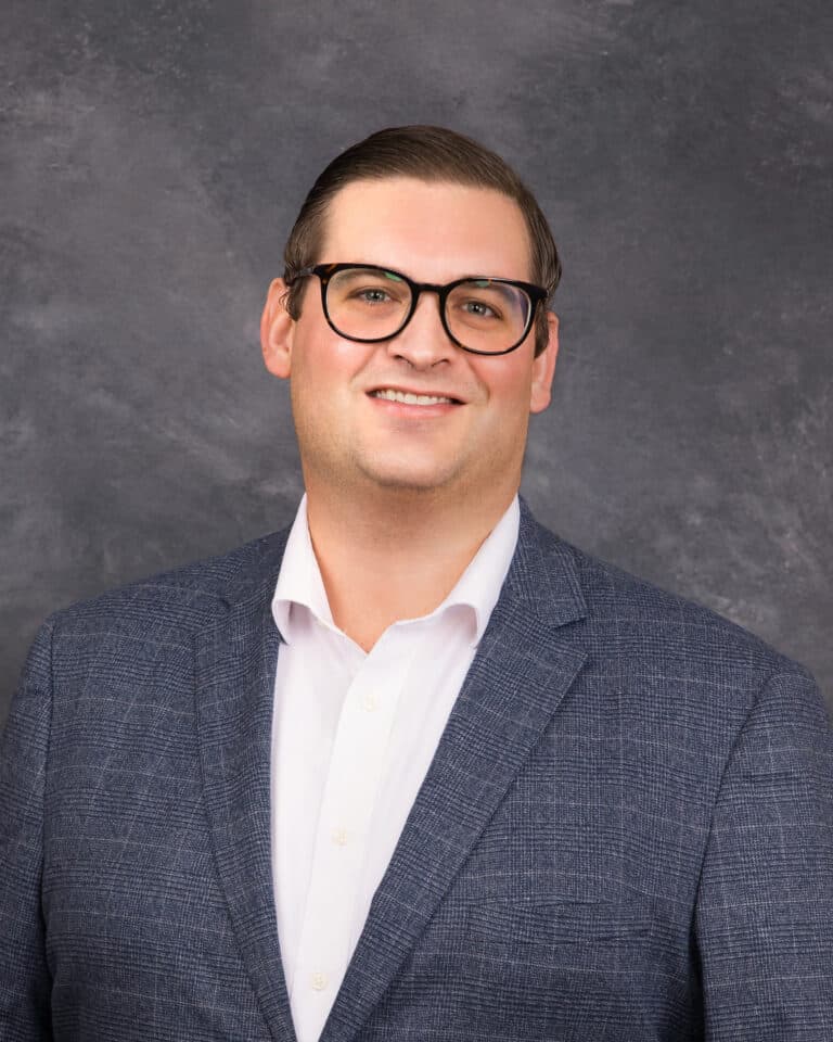 A man with short dark hair, wearing glasses, a white collared shirt, and a blue textured blazer. He is smiling and stands against a grey, mottled background.