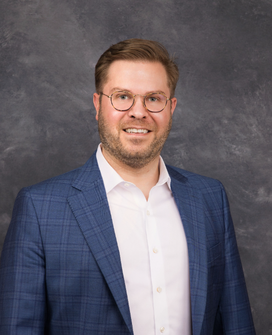 A man with short hair and a beard is wearing glasses, a white shirt, and a blue plaid blazer. He is smiling in front of a gray textured backdrop.