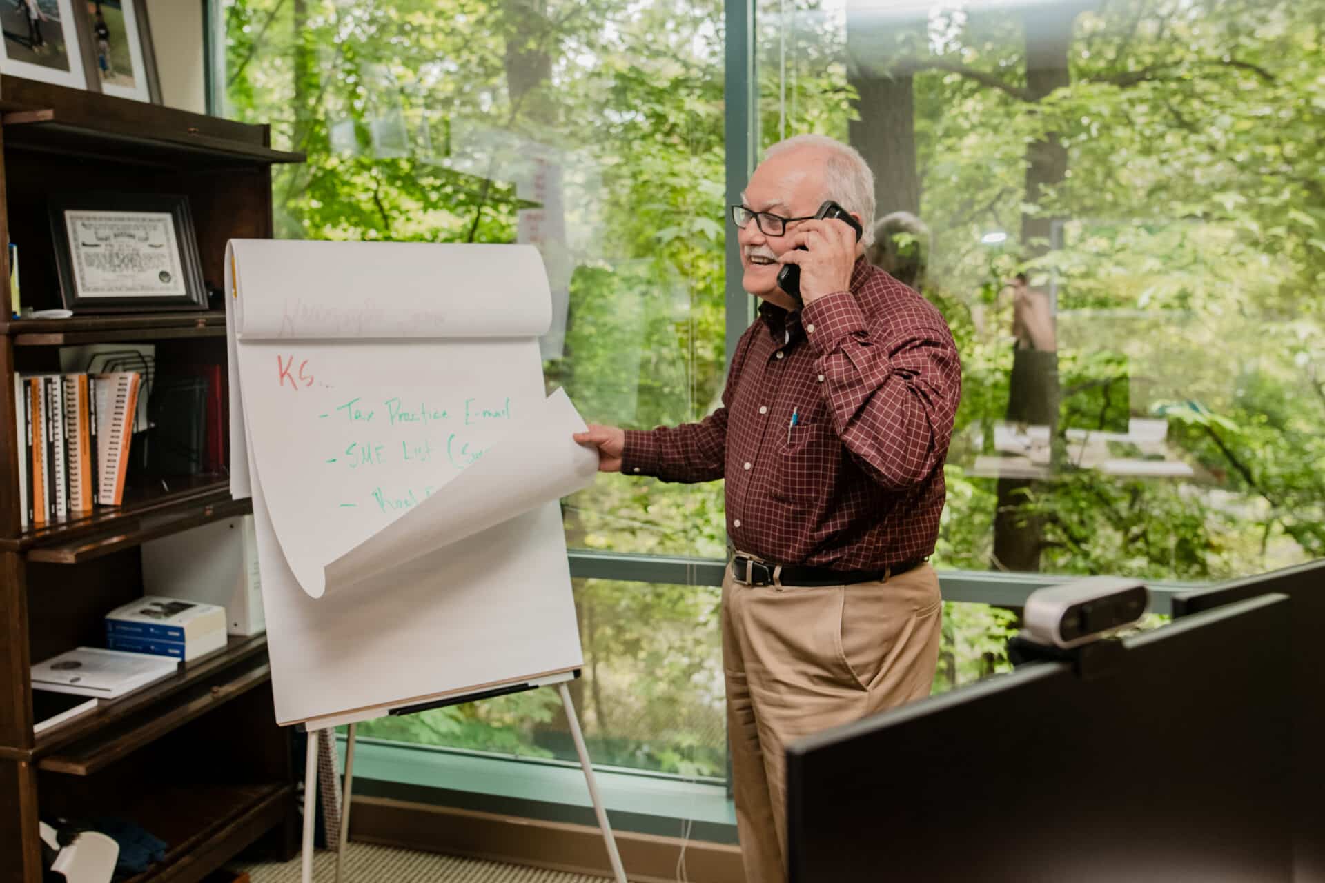 Older man in glasses and plaid shirt on the phone points to a flip chart in a bright office with bookshelves and greenery outside.