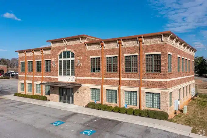 Two-story brick office building with large windows, central entrance, parking lot with handicap spaces, and mostly clear sky above.